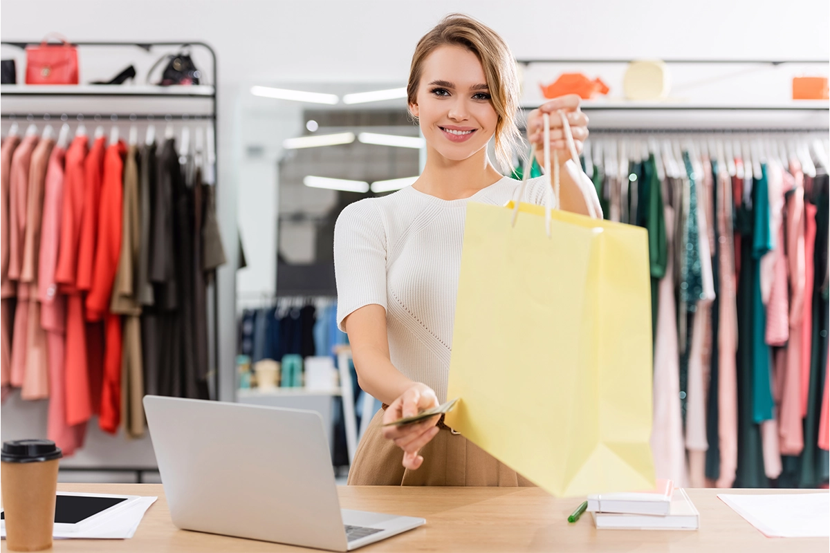 Beautiful female cashier hands customer with shopping bag and card after taking payment.